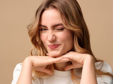 Eine Person mit hellbraunem Haar zwinkert verspielt und stützt ihr Kinn auf die Hände. Sie lächelt leicht und der Hintergrund ist in einem sanften Beige gehalten.