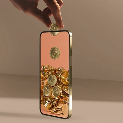 Eine Hand schwebt über einem Smartphone mit hohler Mitte und lässt eine Goldmünze hineinfallen. Das Telefon scheint vor einem braunen, minimalistischen Hintergrund mit ähnlichen Münzen gefüllt zu sein.