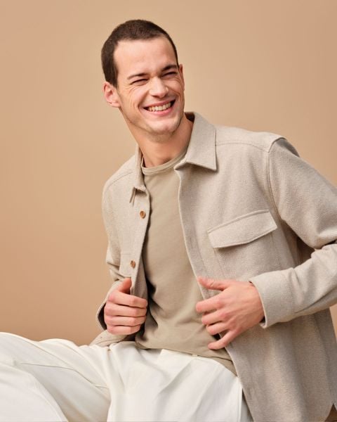 Lächelnder Mann in hellbeiger Jacke über taupefarbenem Hemd und weißer Hose sitzt vor beigem Hintergrund. Er blickt nach links und hält mit den Händen die offene Jacke.