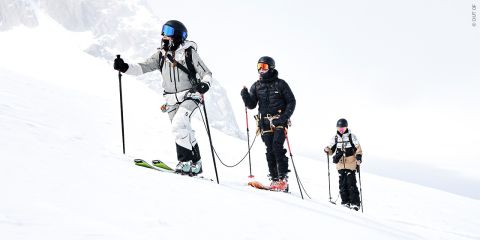 Drei Skifahrer in Winterkleidung, durch Seile verbunden, fahren einen schneebedeckten Hang hinauf. Sie tragen Rucksäcke und Helme mit Schutzbrillen. Im Hintergrund sind ein bewölkter Himmel und eine schneebedeckte Landschaft zu sehen.