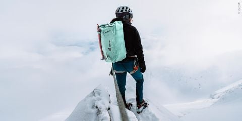 Ein Bergsteiger mit Helm und großem grünen Rucksack steht auf einem schmalen, schneebedeckten Grat. Der Himmel ist bedeckt, Nebel und Wolken verhüllen die Landschaft, was eine dramatische und abenteuerliche Atmosphäre schafft.