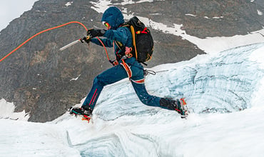 Ein Bergsteiger in blauer Ausrüstung, mit Helm und Eispickel, springt über eine schneebedeckte Gletscherspalte. Gesichert sind sie mit einem orangefarbenen Seil und Spikes an ihren Stiefeln. Im Hintergrund ist ein felsiger Berg zu sehen.