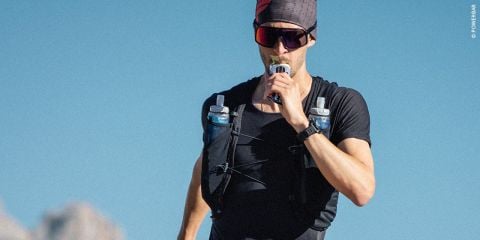 Eine Person in Sportkleidung und Sonnenbrille trinkt aus einem Trinkrucksack mit zusätzlichen Flaschen und steht im Freien unter einem klaren blauen Himmel. Sie trägt einen Hut und eine Armbanduhr.