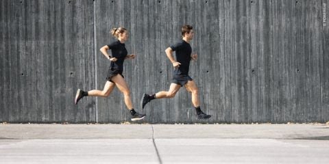 Zwei Personen in Sportkleidung joggen nebeneinander auf einem gepflasterten Gehweg vor einer grauen Betonmauer. Beide tragen schwarze Kleidung und Laufschuhe. Die Szene erinnert an eine urbane Umgebung.