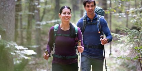 Eine Frau und ein Mann mit Rucksäcken und Wanderstöcken gehen lächelnd auf einem Waldweg spazieren und genießen die Natur inmitten hoher Bäume und des Sonnenlichts, das durch die Blätter fällt.