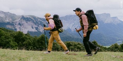 Zwei Personen mit Rucksäcken und Outdoor-Kleidung wandern mit Trekkingstöcken auf einem grasbewachsenen Hügel, im Hintergrund sind Berge und Bäume unter einem bewölkten Himmel zu sehen.