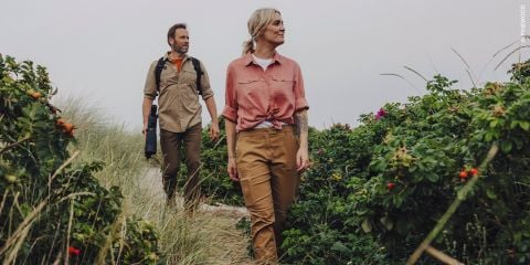 Ein Mann und eine Frau gehen einen Sandweg entlang, der von hohem Gras und grünen Büschen mit Blumen umgeben ist. Beide tragen Straßenkleidung und blicken nach vorne.
