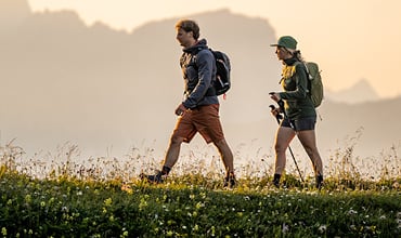 Zwei Menschen mit Rucksäcken und Outdoor-Kleidung wandern durch eine Wiese mit Wildblumen, im Hintergrund sind unter einem warmen, dunstigen Himmel sanft Berge zu erkennen.