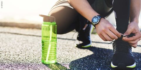 Eine Person bindet ihre schwarzen Laufschuhe auf einer Outdoor-Strecke neben einer grünen Wasserflasche und trägt eine schwarze Uhr am Handgelenk. Im Hintergrund scheint das Sonnenlicht.