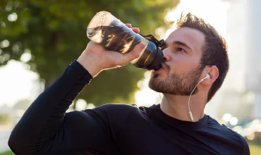 Ein Mann in einem schwarzen Sporthemd trinkt im Freien Wasser aus einer Flasche, während er weiße Kopfhörer trägt, mit Bäumen und Sonnenlicht im Hintergrund.