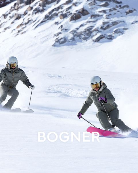 Zwei Skifahrer in bunter Ausrüstung. Links: Person in Gelb mit Schutzbrille, die einen schwarzen Schlitten zieht. Rechts: Zwei Personen in gelben und blauen Jacken, die Skier tragen. Alle tragen Helme und Schutzbrillen. Schneebedeckter Hintergrund mit „BOGNER“ im Text.