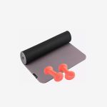 Eine ausgerollte schwarz-graue Yogamatte, auf der zwei kleine orangefarbene Hanteln liegen.