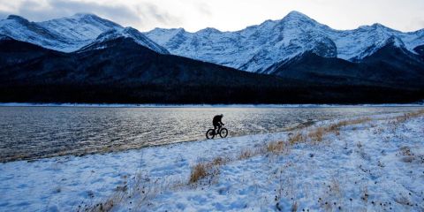 Eine Person fährt mit einem E-Bike auf einem schneebedeckten Weg neben einem See mit Bergen im Hintergrund. Der Text lautet „E-Bike-Tipps für den Winter“.