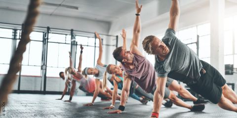 Eine Gruppe von Menschen in Sportkleidung macht während eines Fitnesskurses in einem sonnendurchfluteten Studio seitliche Planks. Der Text „TRAINING NACH DER INNEREN UHR“ ist in fetten weißen Buchstaben darübergelegt.