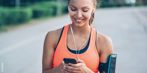 Eine Frau in einem orangefarbenen Tanktop lächelt und schaut auf ihr Smartphone. Sie trägt Kopfhörer und trägt ein Handy am Arm. Der Text „DIE BESTEN FITNESS-APPS“ ist über das Bild gelegt.
