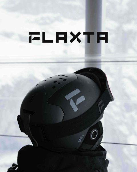Zwei Skifahrer mit Helmen und Ausrüstung stehen auf einer schneebedeckten Metallplattform mit Blick auf eine schneebedeckte Berglandschaft. Sie halten Skier in den Händen und stehen sich gegenüber. Über ihnen prangt in fetten Buchstaben das Wort „FLAXTA“.