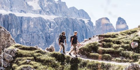 Zwei Menschen wandern auf einem felsigen Bergpfad, umgeben von üppigem grünem Gras und im Hintergrund hoch aufragenden Gipfeln. Der Himmel ist klar und beide Wanderer tragen Rucksäcke. Oben ist der Text „BEINKRAFTTRAINING FÜRS WANDERN“ zu sehen.