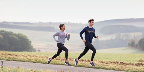 Ein Mann und eine Frau joggen in Sportkleidung auf einem Feldweg. Sie laufen Seite an Seite vor der malerischen Kulisse von Feldern und fernen Hügeln. Der Text lautet: „Gelenke schonen beim Joggen.“.
