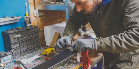 Mann repariert Skier in einer Werkstatt mit Werkzeugen auf einer Werkbank. Der Text lautet „Do It Yourself Ski-Reparatur“. Er trägt Handschuhe und ist auf die Arbeit konzentriert. Verschiedene Geräte und Materialien liegen verstreut herum.