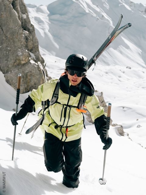 Ein Skifahrer mit Helm, Brille und gelber Jacke klettert einen verschneiten Berghang hinauf, wobei er die Skier auf dem Rücken trägt und die Skistöcke zur Unterstützung benutzt. Felsige Klippen und tiefer Schnee umgeben die Szene.