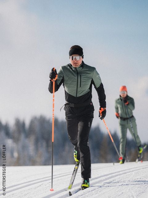 Zwei Skilangläufer auf einer verschneiten Loipe, mit Wintersportausrüstung und Sonnenbrille, mit Bäumen und blauem Himmel im Hintergrund.