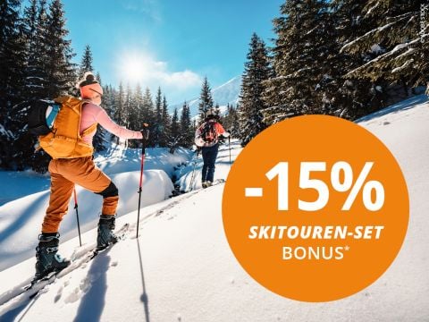 Zwei Skifahrer mit Rucksack und Skistöcken erklimmen einen schneebedeckten Berg. Die sonnige Kulisse zeigt schneebedeckte Gipfel. Ein orangefarbener Kreis markiert ein Angebot „-20% Skitouren-Set Bonus*“.