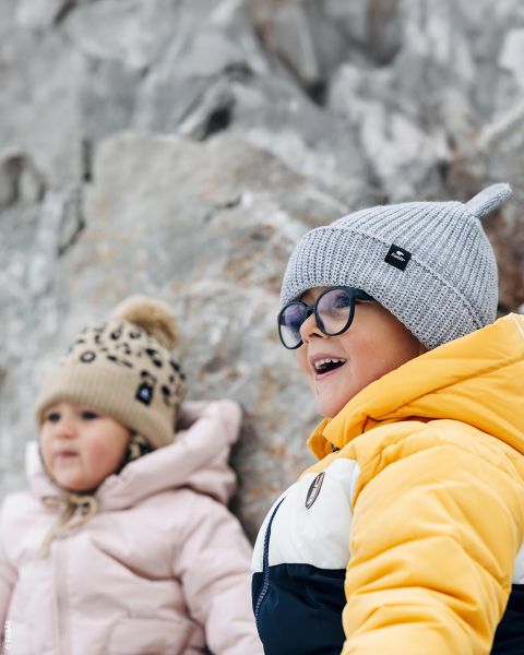 Zwei kleine Kinder in Winterjacken und Strickmützen lächeln im Freien, vor einem felsigen Hintergrund sitzend. Das eine trägt gelb, das andere rosa.