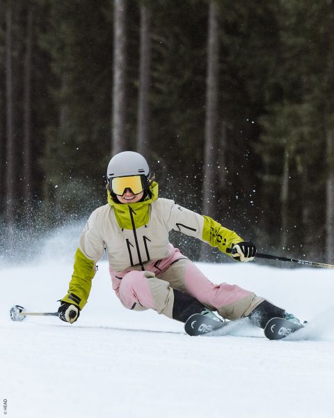Ein Skifahrer mit weißem Helm, gelber Jacke und rosafarbener Hose macht eine scharfe Kurve auf einer verschneiten Piste, wobei der Schnee in die Luft gespritzt wird.