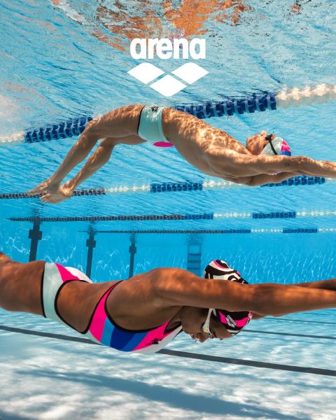 Ein Schwimmer mit schwarzer Schwimmkappe und reflektierender Schwimmbrille gleitet unter Wasser in einem mit Bahntrennwänden markierten Becken. Der blaue Badeanzug ist mit orangefarbenen Verzierungen versehen. Das Wasser ist klar, und darüber ist das Arena-Logo zu sehen.