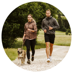 Zwei Personen joggen auf einem Kiesweg in einem grünen Park, neben ihnen ein angeleinter Hund. Sie tragen sportliche Kleidung und schauen nach vorne.