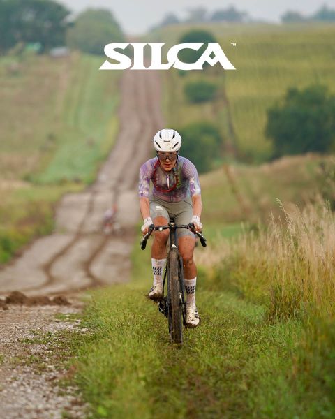 Radfahrer, der auf einer Schotterstraße bergauf durch grasbewachsene Felder fährt. Er trägt einen Helm und eine Radfahrerkleidung, mit dem "SILCA"-Logo am oberen Bildrand.