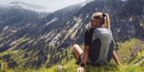 Eine Frau mit einem Rucksack sitzt auf einem grasbewachsenen Hügel und blickt auf eine malerische Berglandschaft mit Wäldern und Felsen.