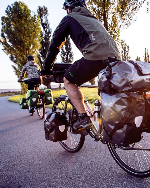 Zwei Radfahrer mit Gepäcktaschen fahren auf einer gepflasterten Straße, die von Bäumen umgeben ist und in warmes Sonnenlicht getaucht wird.