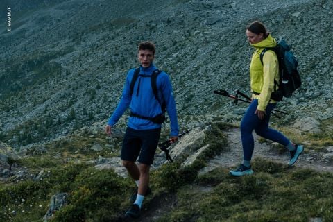 Zwei Personen in Outdoor-Kleidung wandern auf einem felsigen Bergpfad mit Trekkingstöcken durch das zerklüftete Gelände und die grüne Landschaft im Hintergrund.