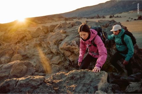 Zwei Wanderer in bunten Jacken klettern bei Sonnenaufgang über felsiges Gelände, während das Sonnenlicht über die zerklüftete Landschaft im Hintergrund fällt.