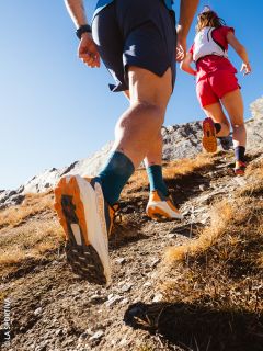Zwei Trailrunner, von hinten gesehen aus einem niedrigen Winkel, laufen auf einem felsigen Pfad unter einem klaren blauen Himmel bergauf.