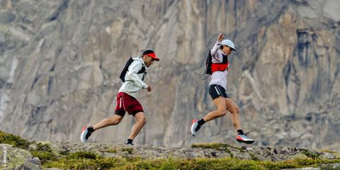 Zwei Personen beim Trailrunning in felsigem Gelände mit einer großen Felswand im Hintergrund, beide mit Rucksäcken, Mützen und Sportkleidung.