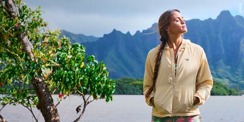 Eine Frau in einer beigen Jacke steht an einem See mit Bergen im Hintergrund, die Augen geschlossen und das Gesicht der Sonne zugewandt, neben einem belaubten Baum.
