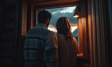 Ein Paar in kuscheligen Pullovern steht an einem Hüttenfenster und beobachtet gemeinsam einen Sonnenuntergang in den Bergen bei warmem Licht.