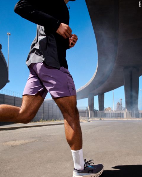 Eine Person läuft an einem sonnigen Tag im Freien unter einer großen Überführung, trägt lila Shorts, ein schwarzes Hemd, weiße Socken und Laufschuhe.