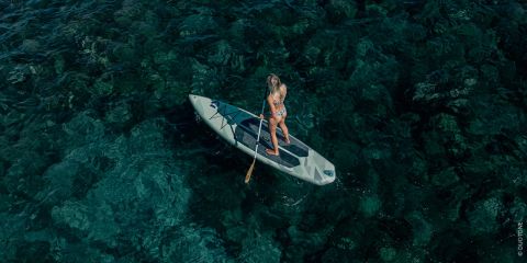 Eine Frau im Bikini paddelt auf einem Stand-up-Paddleboard über klares, tiefblaues Wasser mit sichtbaren Felsen unter der Wasseroberfläche.