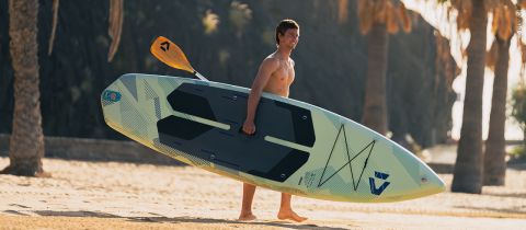 Mann ohne Hemd, der am Sandstrand spazieren geht und ein Stand-up-Paddleboard und ein Paddel trägt, mit Palmen im Hintergrund.