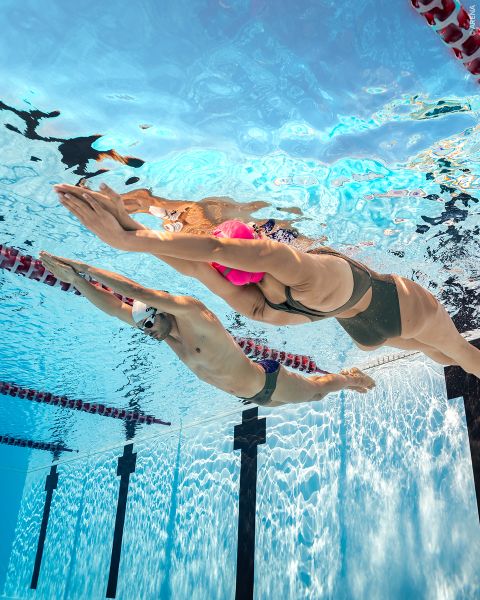 Zwei Schwimmer, von denen einer eine rosafarbene und der andere eine weiße Kappe trägt, gleiten in einem Schwimmbad Seite an Seite unter Wasser, wobei sie in getrennten Bahnen schwimmen.