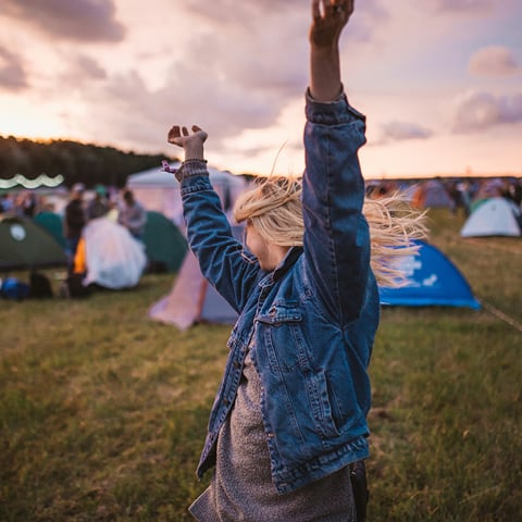 Eine Person in Jeansjacke hebt freudig die Arme auf einem Campingplatz mit Zelten bei Sonnenuntergang.