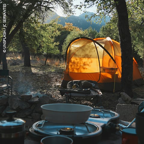 Ein leuchtend orangefarbenes Zelt auf einem bewaldeten Campingplatz mit Kochgeschirr und Campingzubehör im Vordergrund.