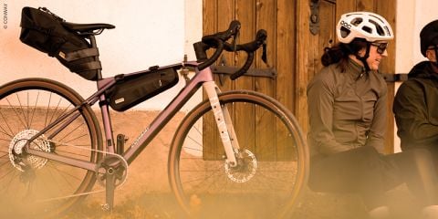 Zwei Radfahrer, die Helme und Jacken tragen, sitzen auf dem Boden in der Nähe einer Mauer; neben ihnen steht ein voll beladenes Tourenrad.