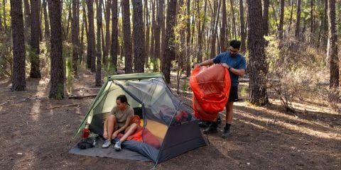 Zwei Menschen, die im Wald zelten; einer sitzt im Zelt, während der andere draußen steht und einen orangefarbenen Schlafsack in der Hand hält.