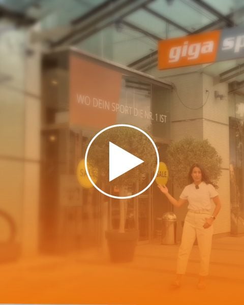 Una mujer vestida de blanco está de pie frente a una tienda con un letrero de "Giga" y una pancarta que dice "DONDE TU DEPORTE ES EL NÚMERO 1", haciendo un gesto con una mano.