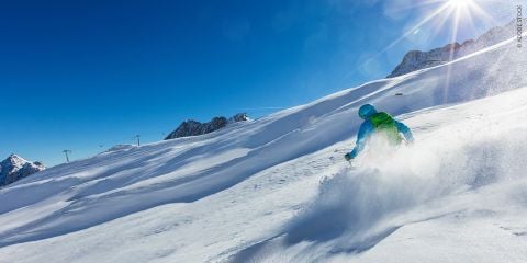 Ein Skifahrer in roter Hose und blauer Jacke fährt unter strahlend blauem Himmel einen schneebedeckten Hang hinunter. Die Sonne scheint hell und beleuchtet den Schnee. In der oberen linken Ecke steht „Winter Sport Services“.