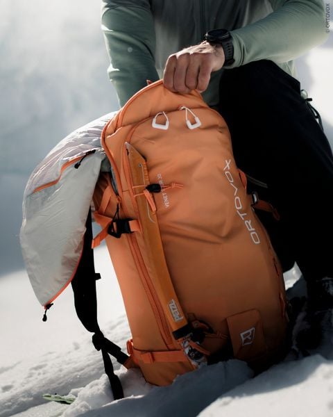 Ein Wanderer öffnet einen orangefarbenen Ortovox-Rucksack in einer verschneiten Berglandschaft.